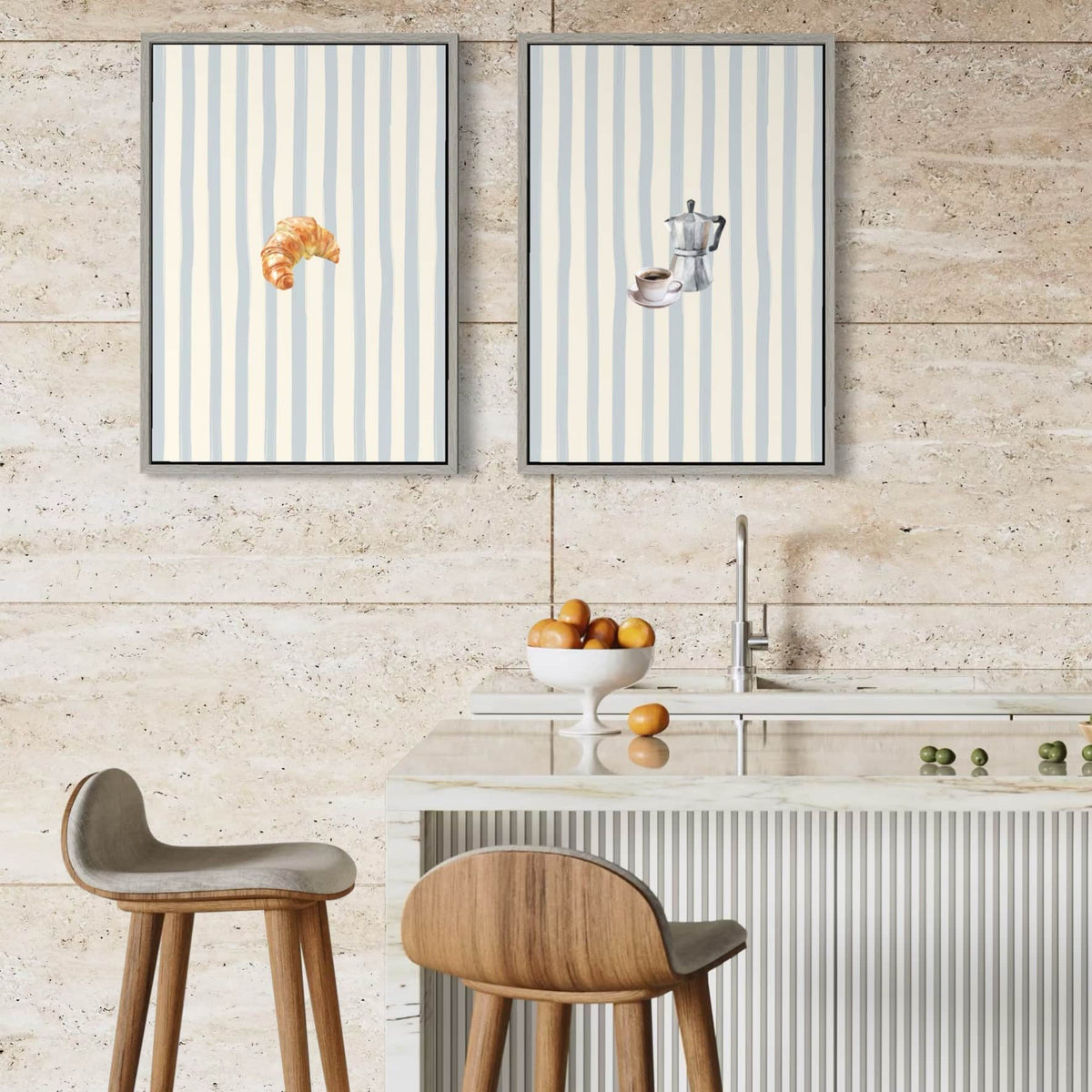 Two vertical framed Solstice Studio wall art prints with pale blue and white stripes hang above a kitchen island. The left print shows a croissant, and the right print shows a coffee cup and saucer.