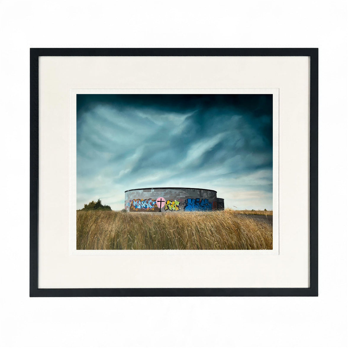 Framed landscape art print  "Vessel" by Zoe Marsden, featuring a circular concrete structure covered in colourful graffiti, situated in a field of dry grass under a dramatic cloudy sky. 