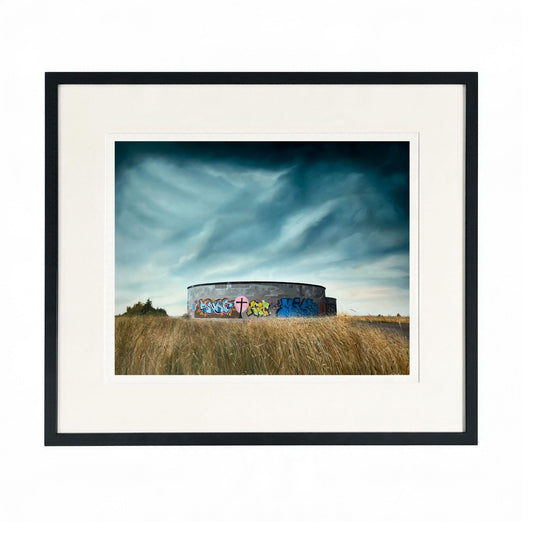 Framed landscape art print  "Vessel" by Zoe Marsden, featuring a circular concrete structure covered in colourful graffiti, situated in a field of dry grass under a dramatic cloudy sky. 
