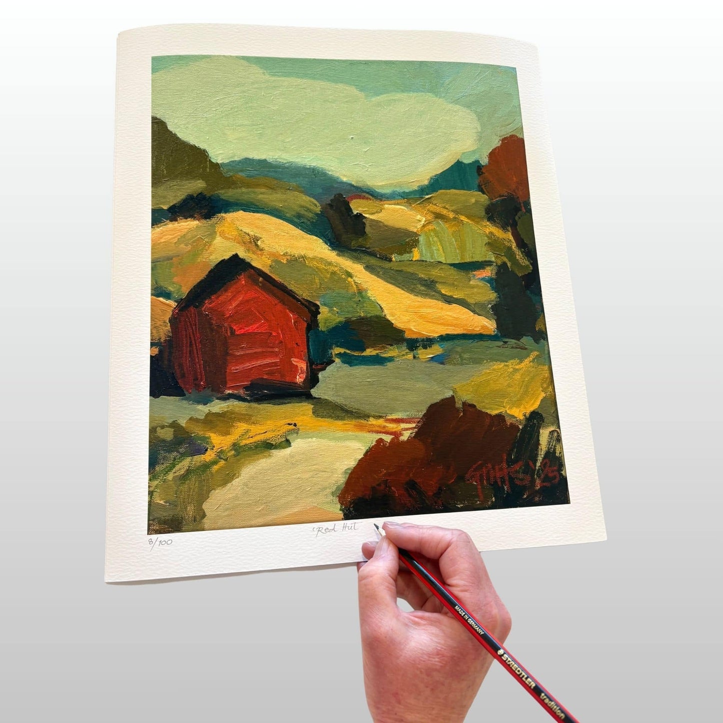 Hand holding a pencil to sign a print of a colourful landscape with a red barn.