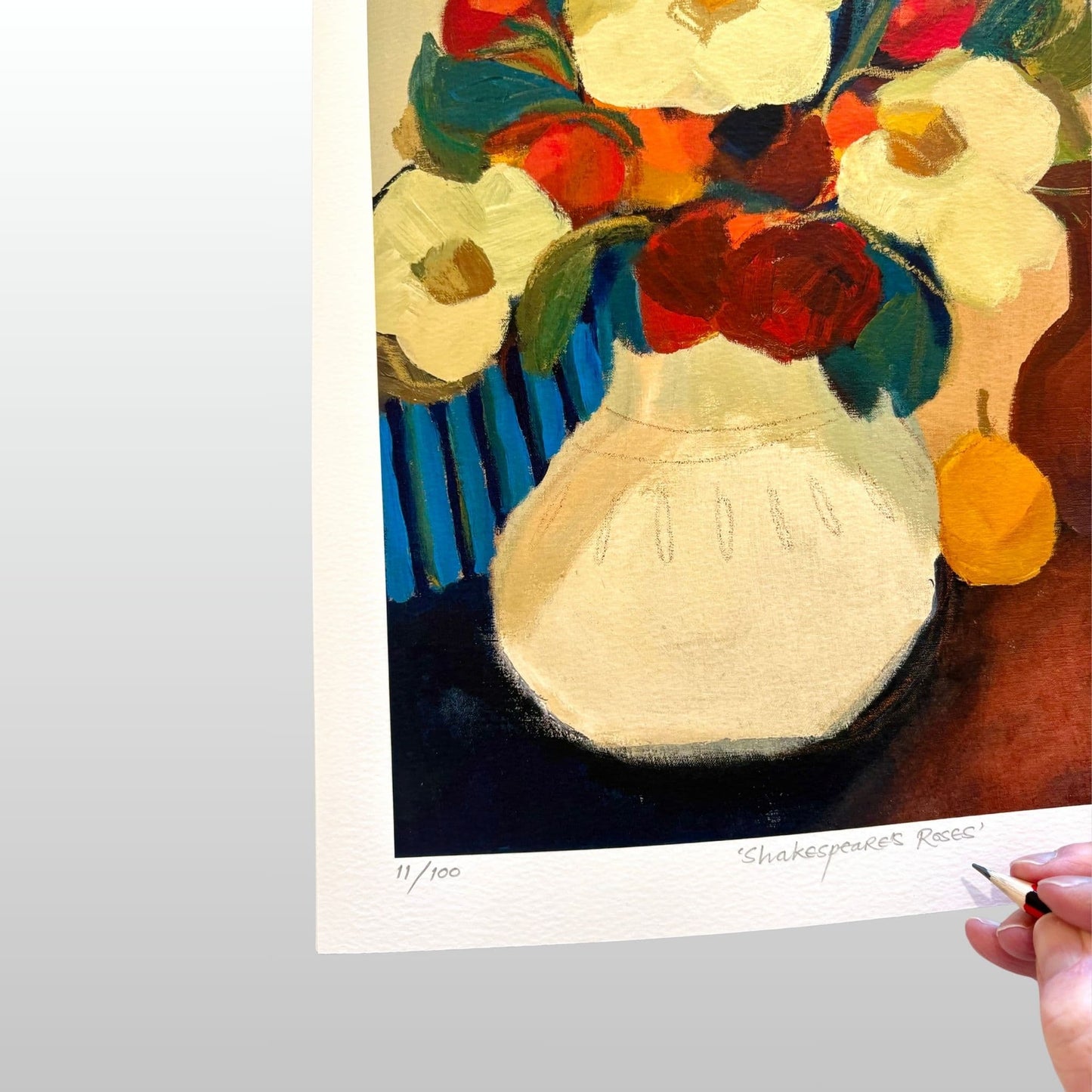 Art print of a colourful still life with flowers and a vase, being signed by a hand holding a pencil.