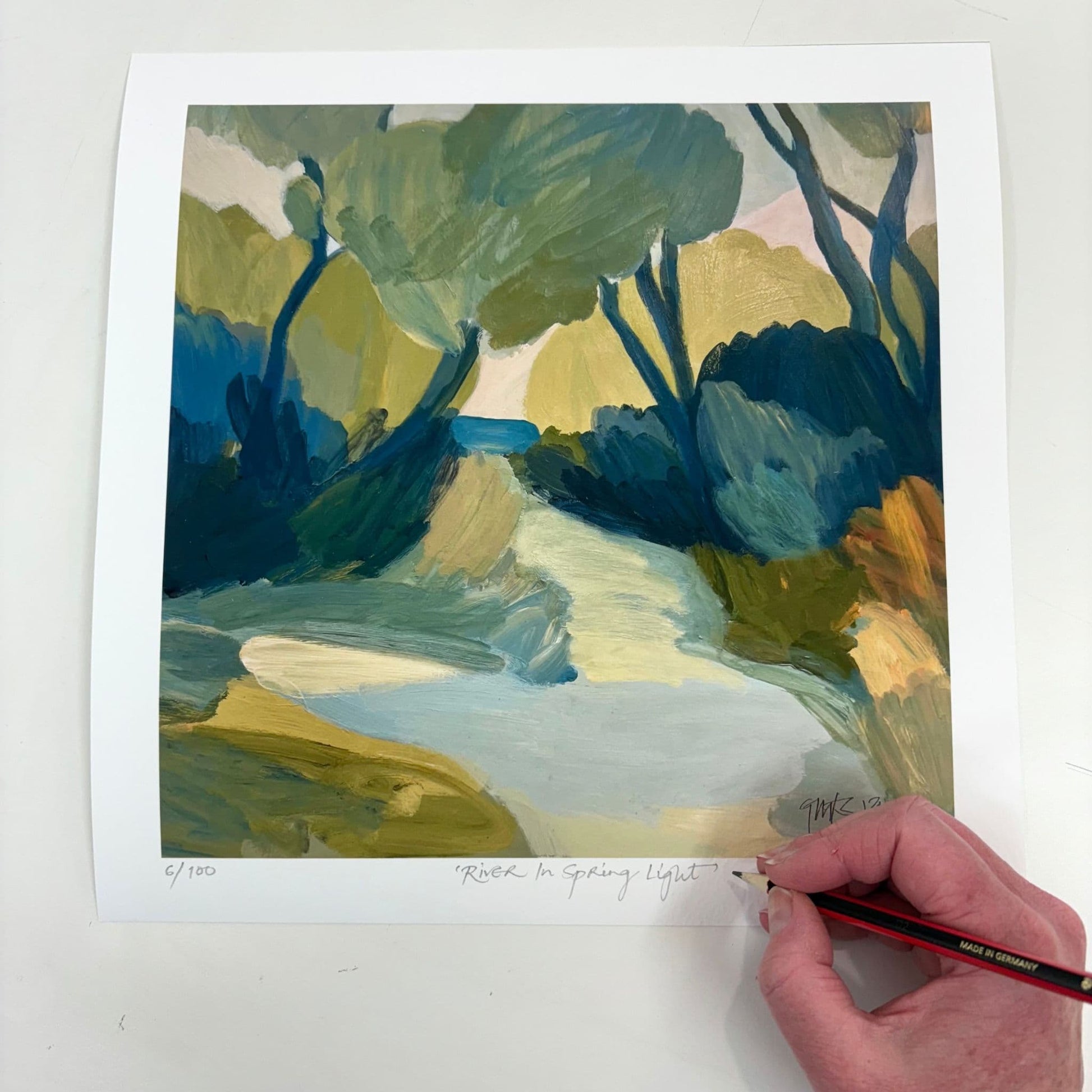 Hand signing Georgina Hoby Scutt's "River in Spring Light" limited edition print – A vibrant river scene where light and shadow dance across rich blues. 