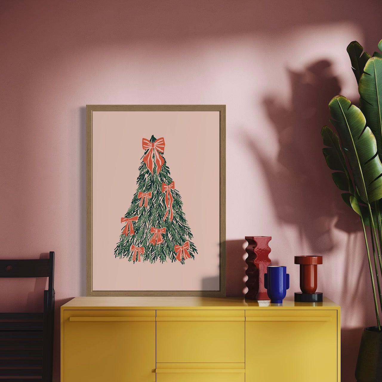 Framed artwork of a Christmas tree with red bows on a pink wall above a yellow cabinet.