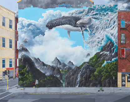 Mural of a whale emerging from water with mountains and a person in the foreground on a city street.