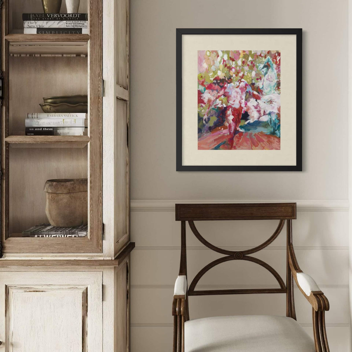 A framed limited edition print, Fall Out, by Justine Turnbull with pink, red, green, and blue brushstrokes, displayed on a light gray wall above a wooden chair next to a rustic cabinet.