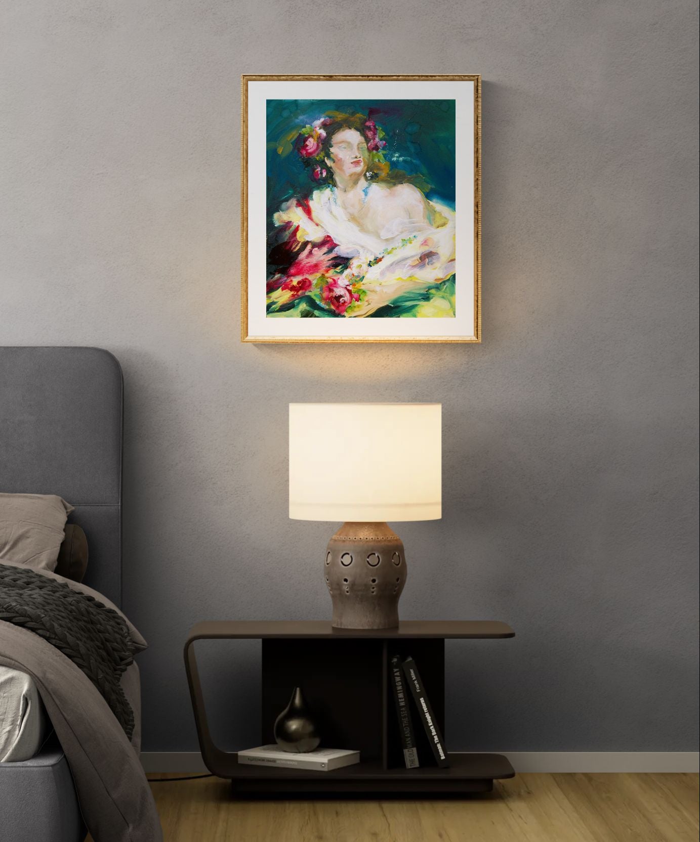 A framed Anita DeSoto portrait hangs on a grey wall above a bedside table with a lit lamp and books, next to a bed.