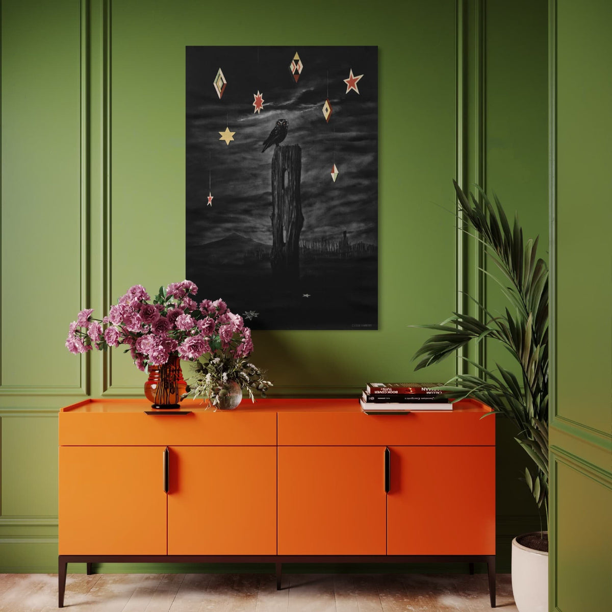 Hanging on a green wall, Nocturnal by Paula Coulthard, showing a black ruru perched on a weathered wooden post against a cloudy night sky, with hanging geometric and star-shaped ornaments in cream, red, and gold.