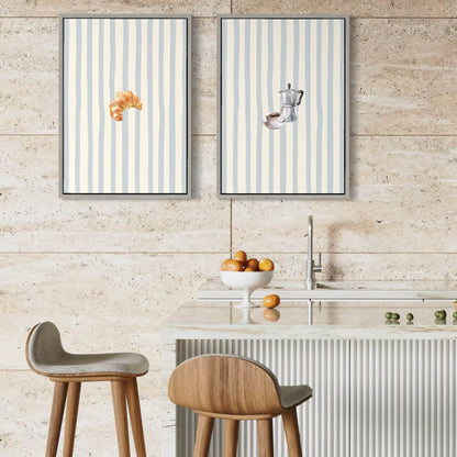 Two vertical framed Solstice Studio wall art prints with pale blue and white stripes hang above a kitchen island. The left print shows a croissant, and the right print shows a coffee cup and saucer.