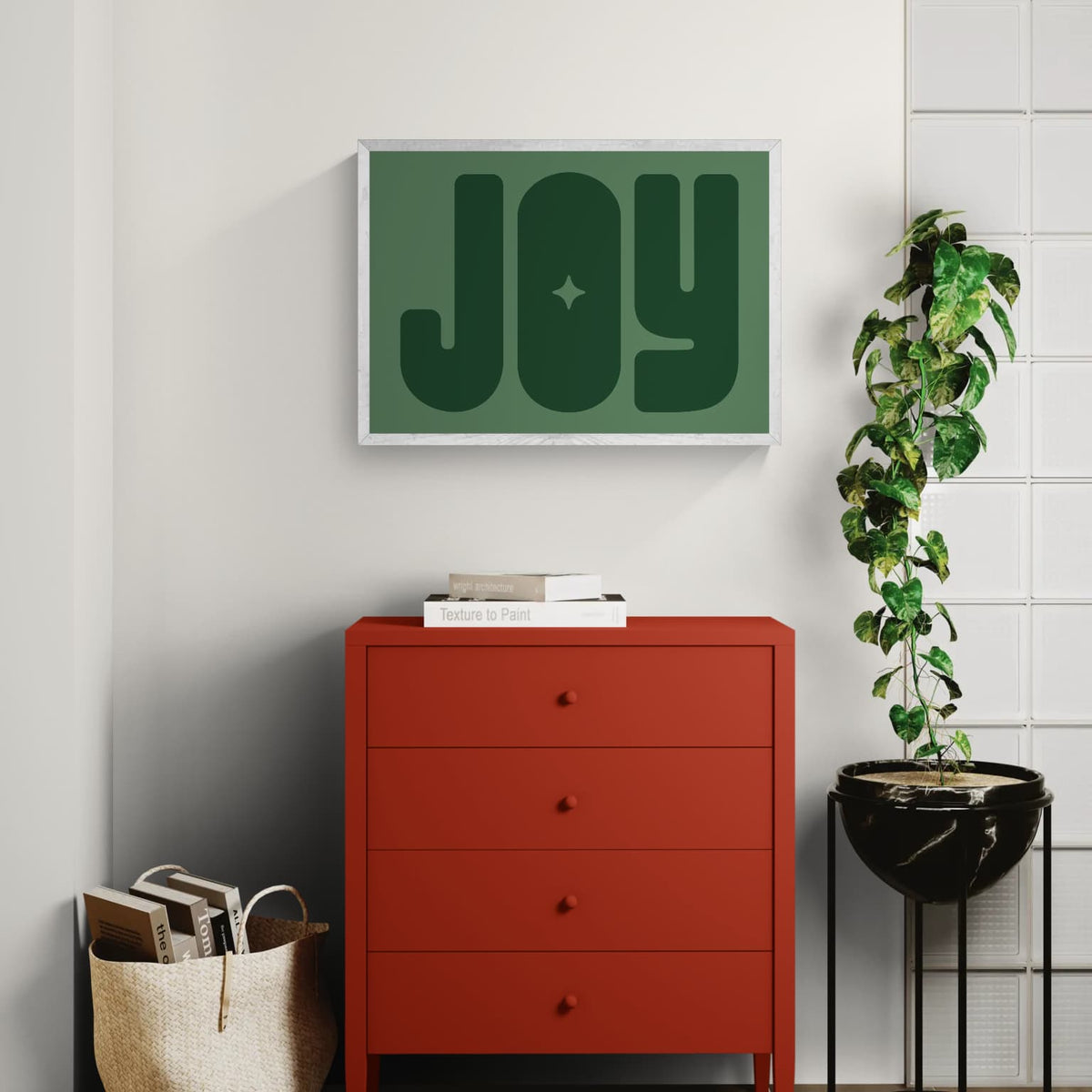 A framed wall art print by Solstice Studio Prints, featuring the word 'JOY' in dark green on a muted sage green background, displayed above an orange chest of drawers. 