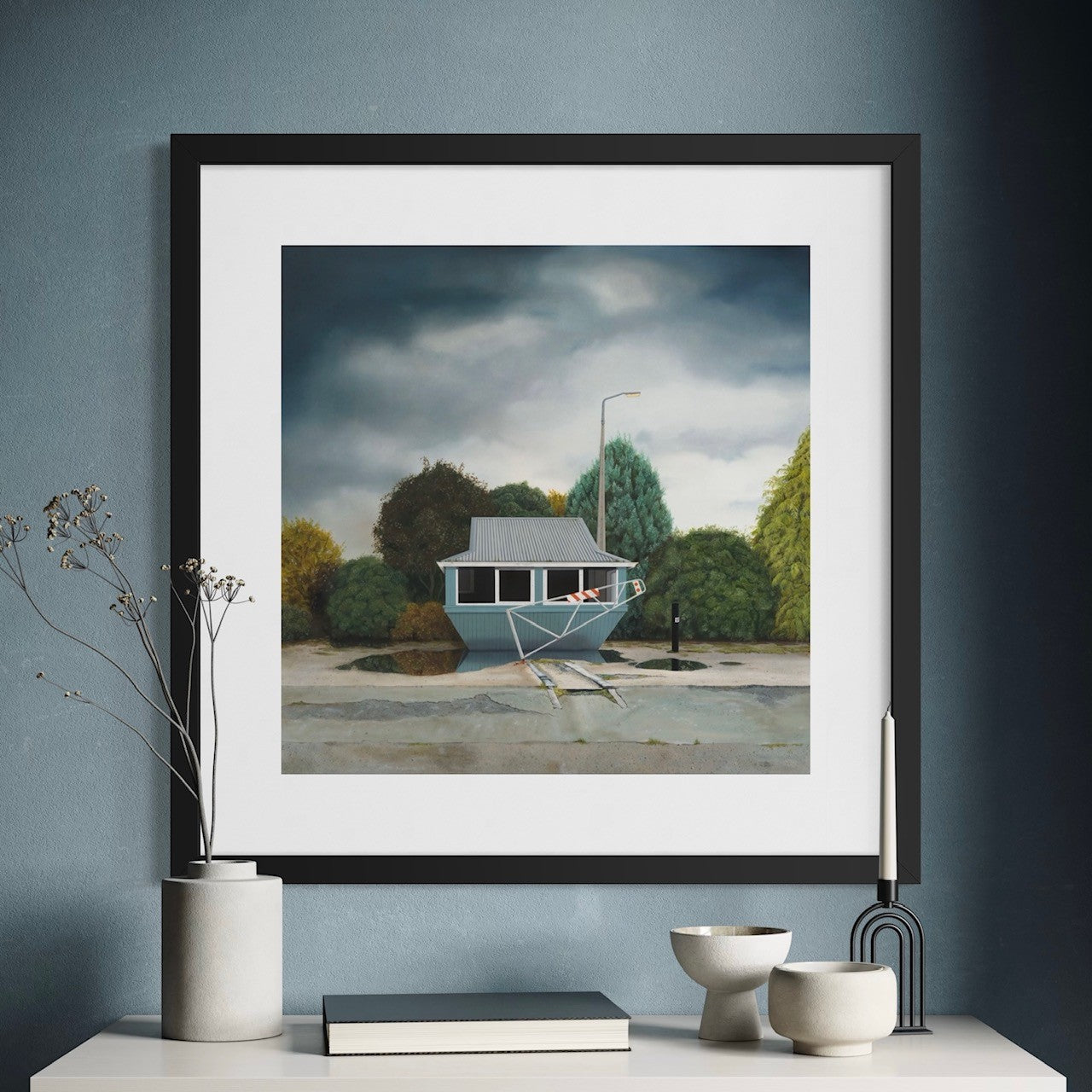 Framed artwork of an abandoned building with green trees and a moody sky, hanging on a blue wall with decorative items below.