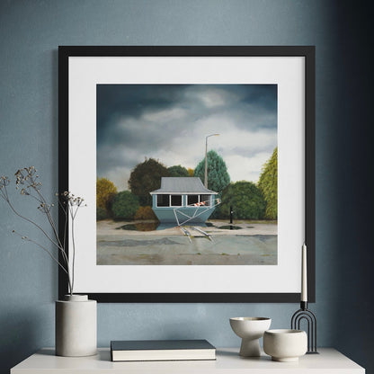 Framed artwork of an abandoned building with green trees and a moody sky, hanging on a blue wall with decorative items below.