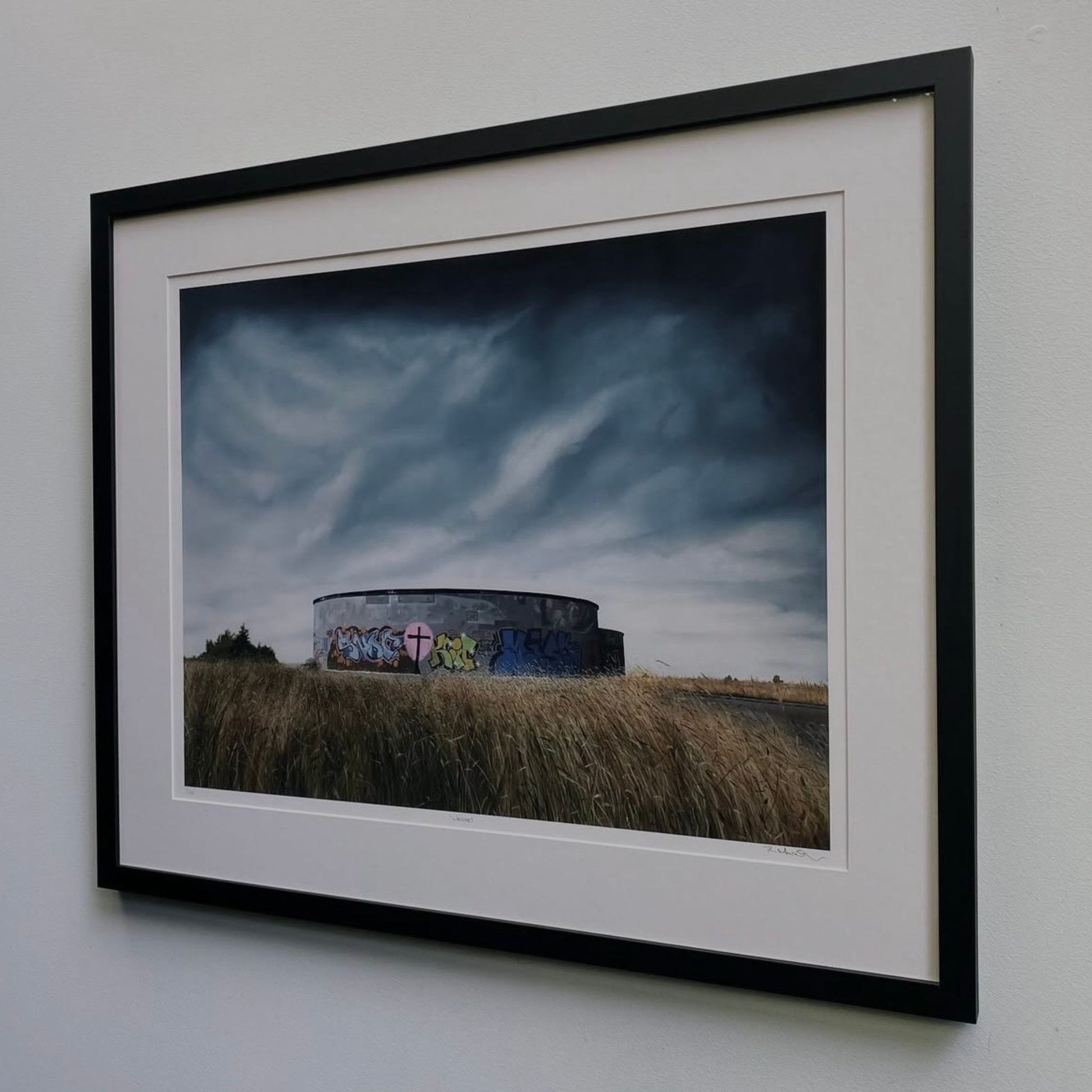 Framed and hanging print of a round structure with graffiti against a dramatic sky.