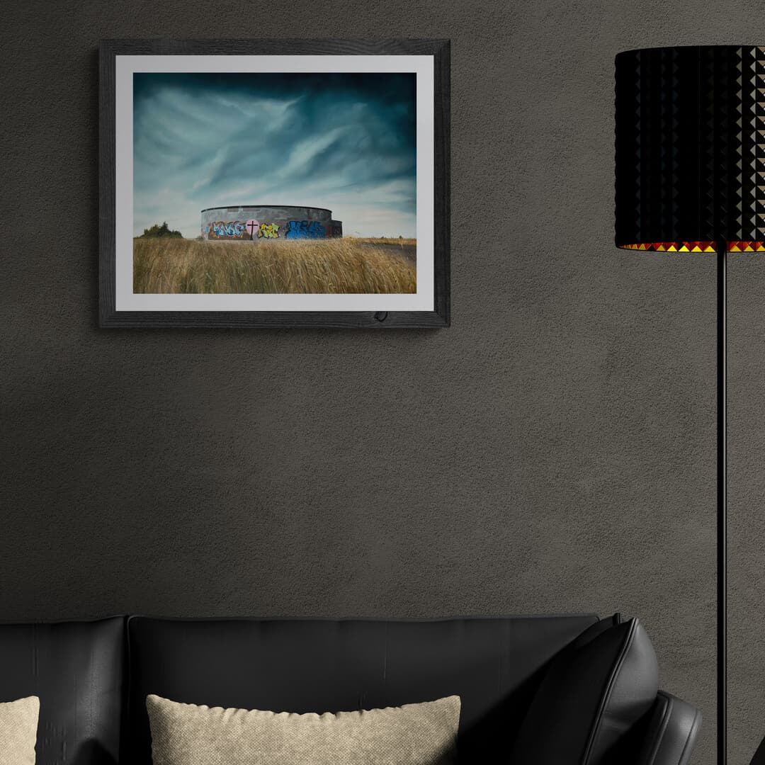 Framed artwork of a landscape with a concrete building on a gray wall above a black sofa.
